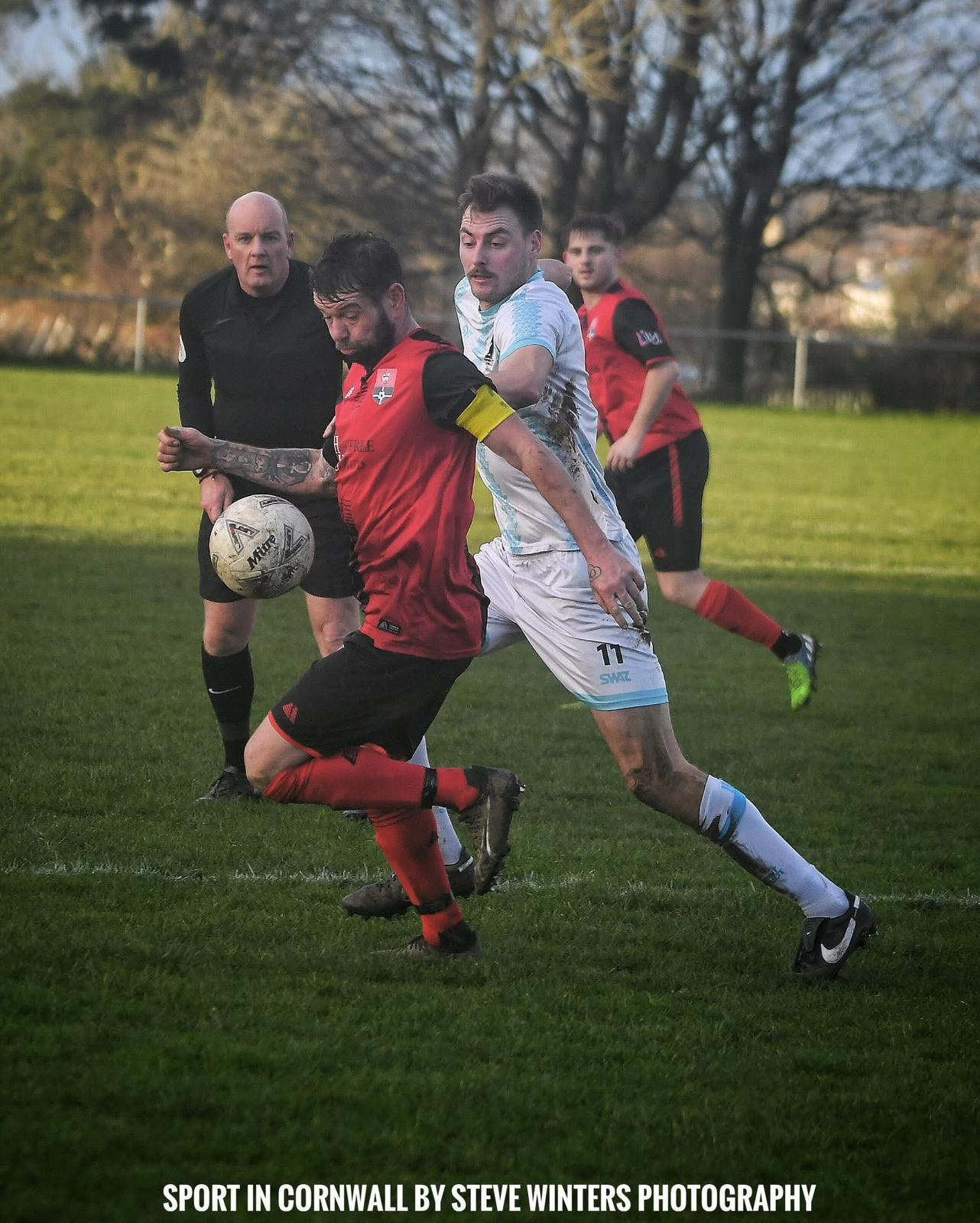 Redruth United men's player in red and black shielding the ball from an opponent while the referee watches